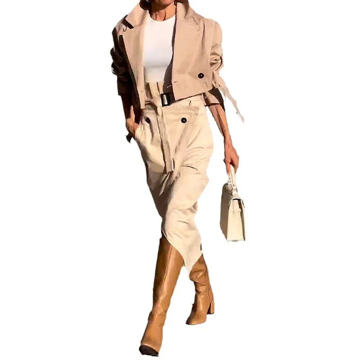 Woman in beige cropped jacket, high-waisted skirt, tan knee-high boots, and white handbag on white background