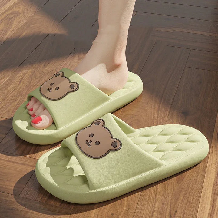Light green cushioned slide slippers with brown bear face design worn on wooden floor