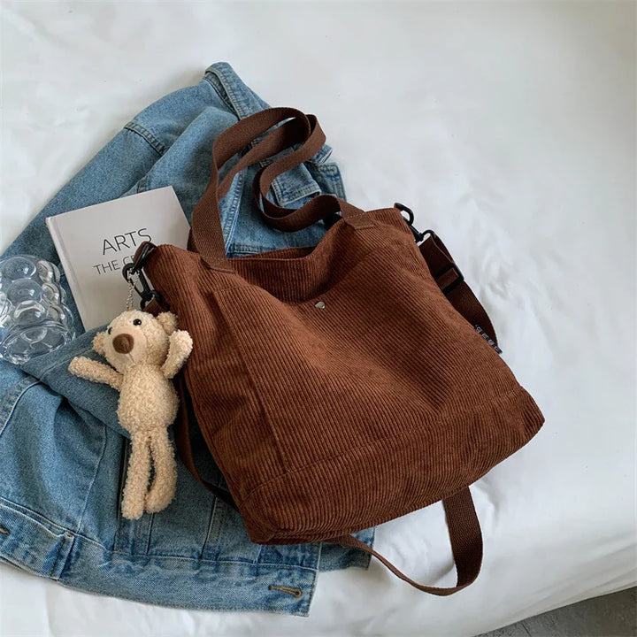 Brown corduroy shoulder bag with teddy bear keychain on denim jacket and book on white bed