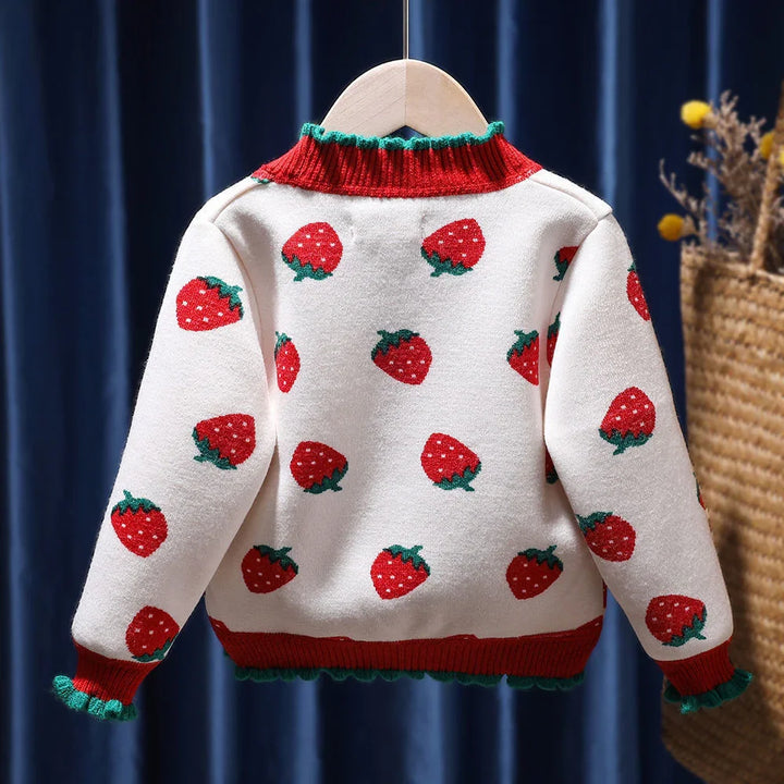 White knit sweater with red strawberry pattern, red collar and cuffs with green trim hanging on wooden hanger