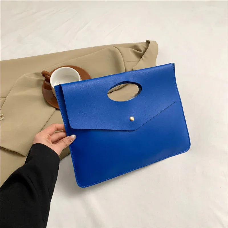 Hand holding a blue envelope clutch bag with a cutout handle, beige jacket and white cup on saucer in background