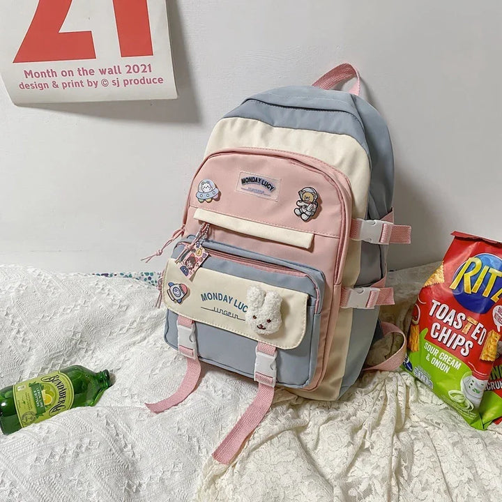 Pastel pink, gray, and cream backpack with cute patches and bunny keychain on white textured blanket, snack bag nearby