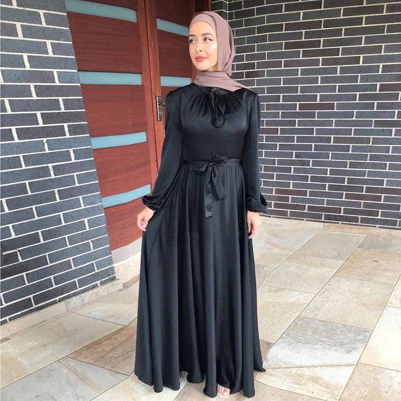 Woman in a black long-sleeve maxi dress with waist tie and mauve hijab standing by brick wall