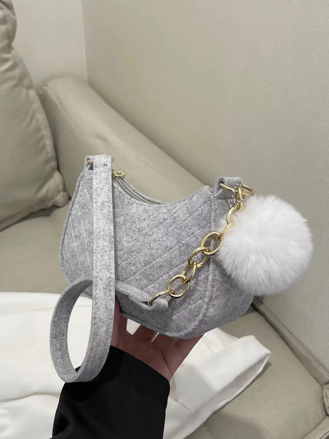 Gray quilted felt handbag with gold chain and white fluffy pom-pom keychain on beige couch
