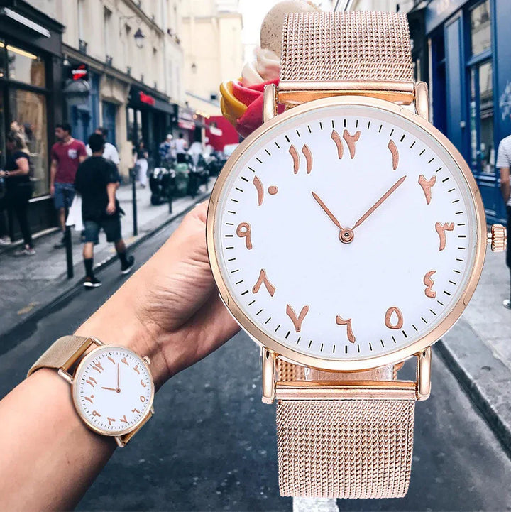 Hand wearing rose gold mesh strap watch with Arabic numerals on white dial on city street