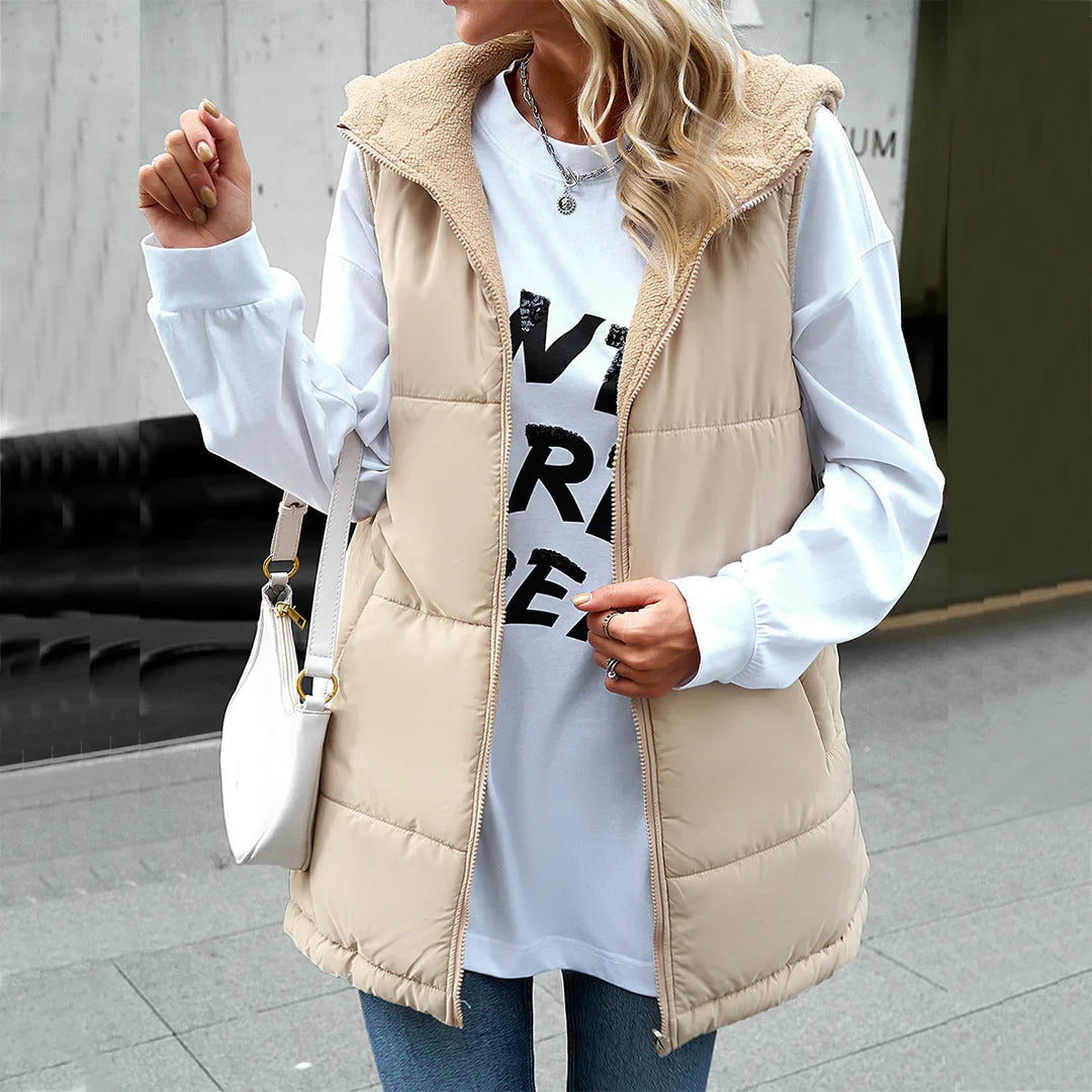 Woman wearing beige hooded puffer vest over white long sleeve shirt with black print, carrying white handbag