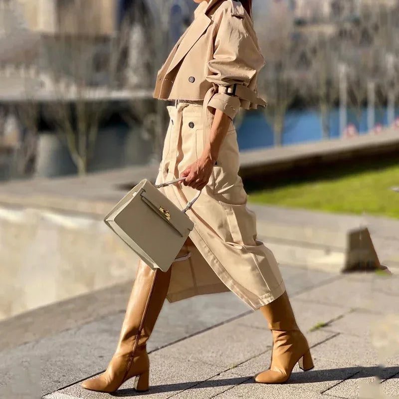 Woman walking outdoors in tan trench coat and boots, carrying beige handbag on city sidewalk