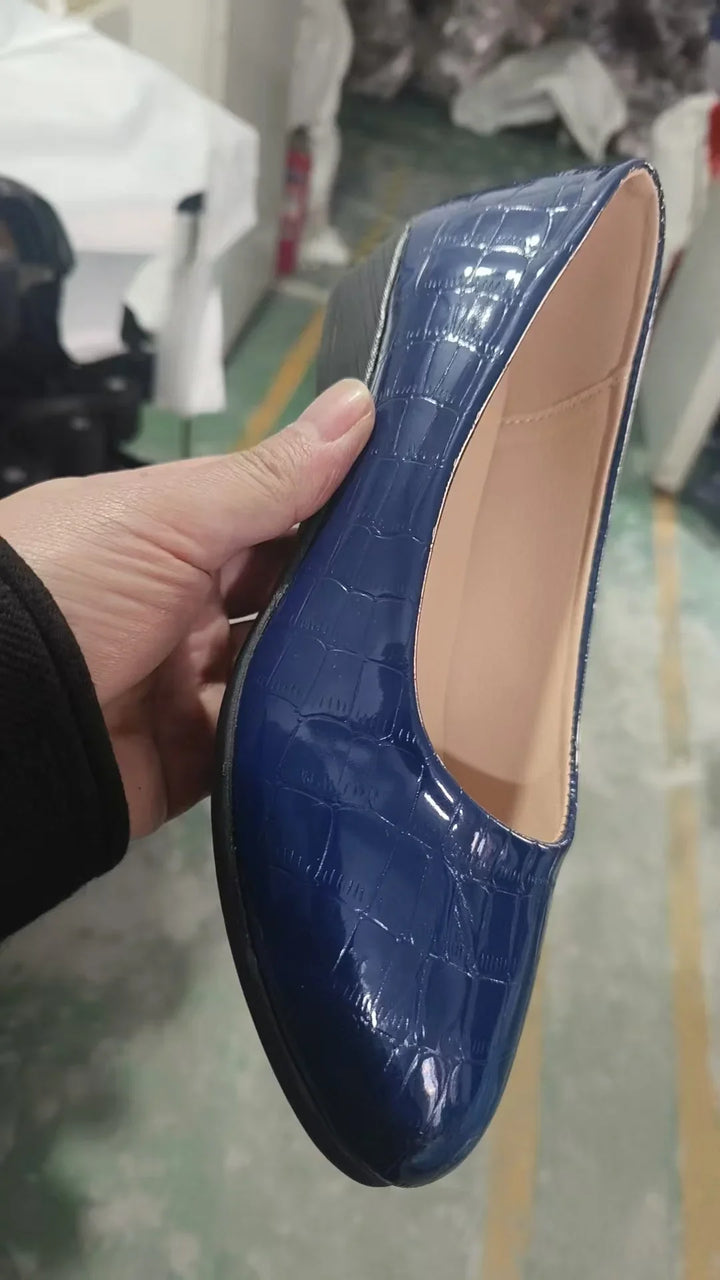 Hand holding a glossy blue crocodile pattern flat shoe with beige interior in a factory setting