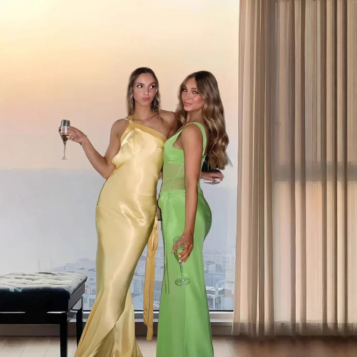 Two women wearing elegant satin dresses in yellow and green holding wine glasses indoors
