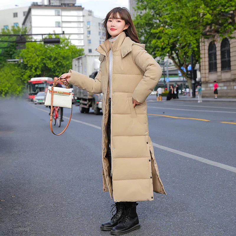 Woman standing on city street wearing long beige puffer coat and black boots, holding white handbag