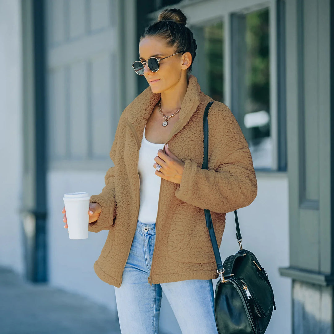 Woman wearing tan sherpa jacket, white tank top, jeans, sunglasses, holding coffee cup with black handbag