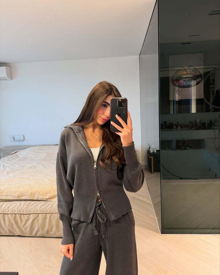 Young woman in dark gray loungewear taking a mirror selfie in modern bedroom with beige bed and glass shower
