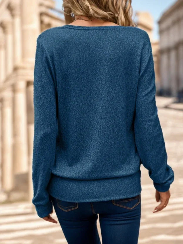 Woman wearing a blue long-sleeve knit sweater and dark jeans standing outdoors with blurred architectural background