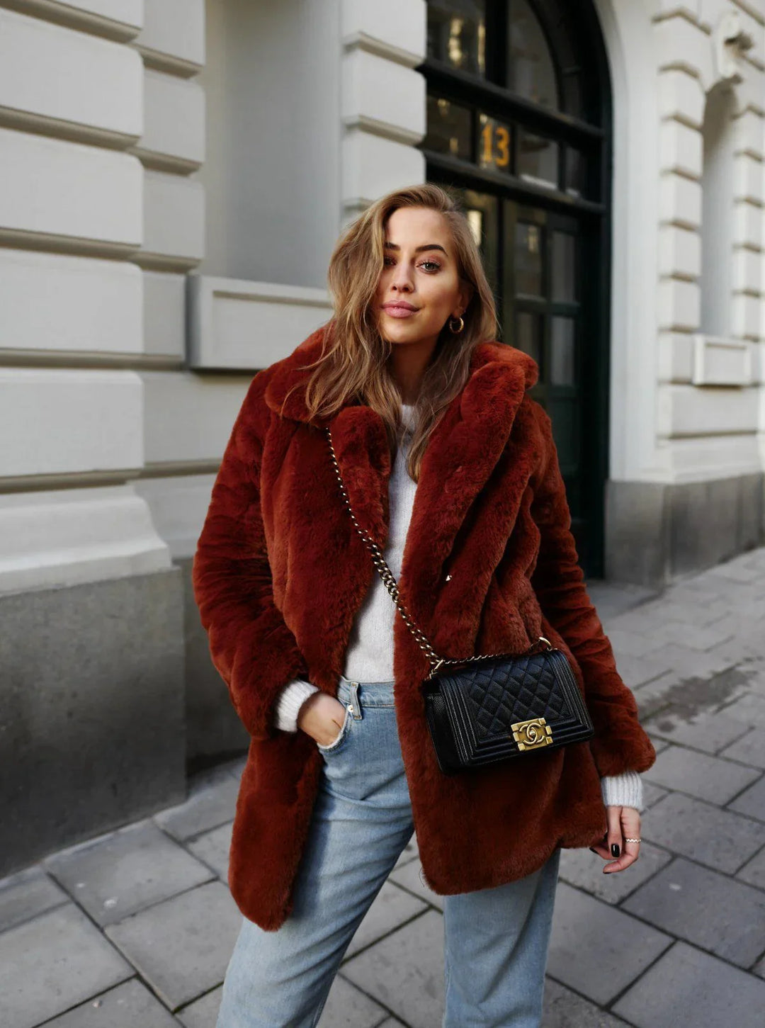 Woman wearing rust-colored faux fur coat, light sweater, jeans, black quilted purse standing on city sidewalk