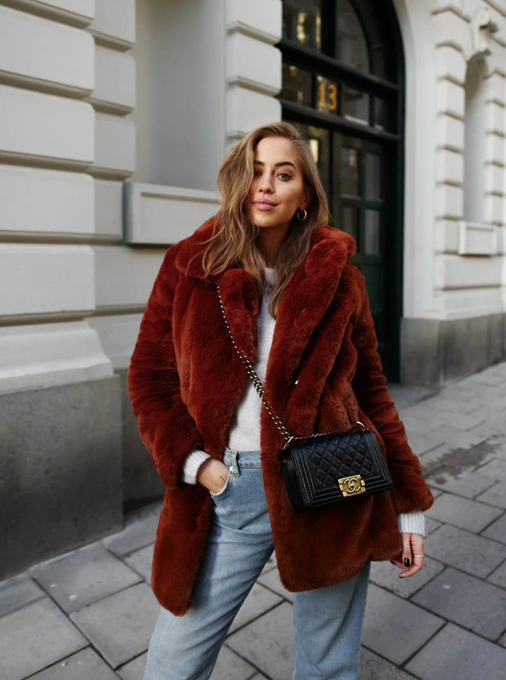 Woman wearing rust-colored faux fur coat, light sweater, jeans, black quilted purse standing on city sidewalk