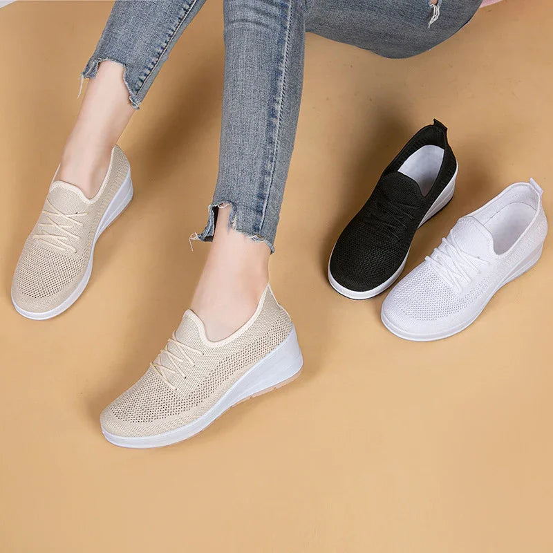 Woman wearing beige knitted slip-on sneakers with white sole, next to black and white sneaker pairs on tan background
