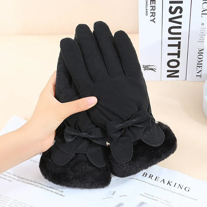 Black winter gloves with fur cuffs and bow details, held by hand on a beige surface