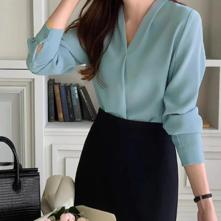Woman wearing a light blue blouse and black skirt standing indoors with a black handbag and bouquet