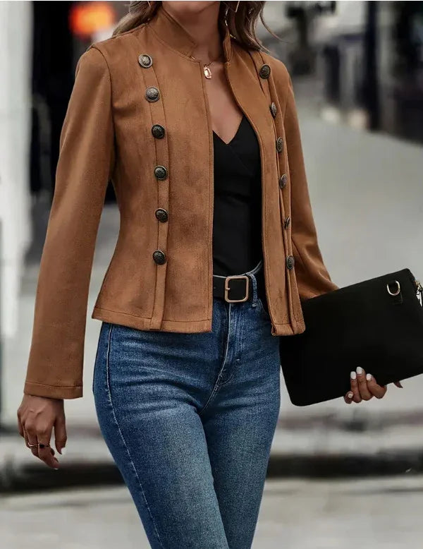 Woman wearing brown buttoned military-style jacket with black top, blue jeans, and holding black clutch