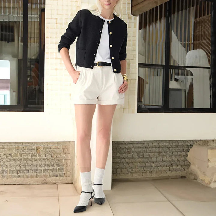 Woman wearing black cardigan, white top, cream shorts, black belt, white socks, and black heels standing indoors