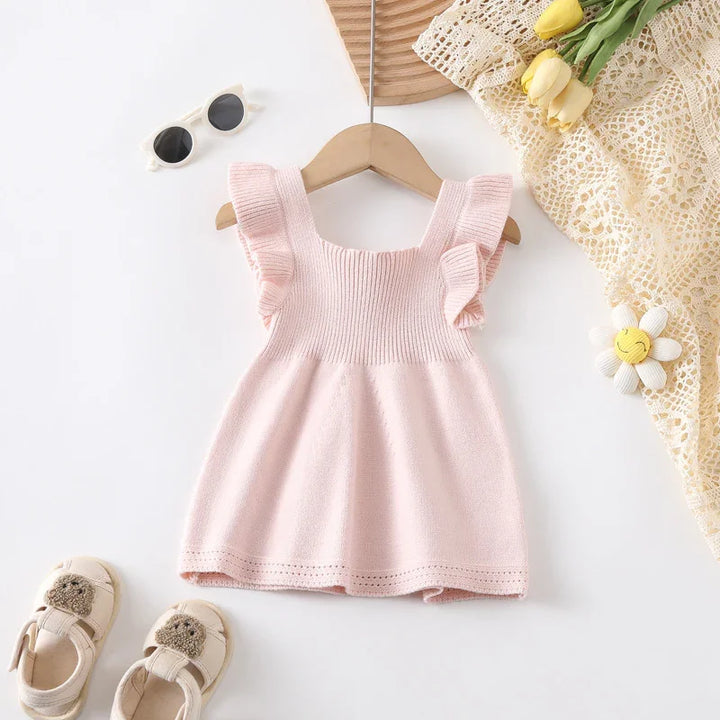 Light pink knitted baby dress with ruffled straps on hanger, paired with beige shoes and sunglasses
