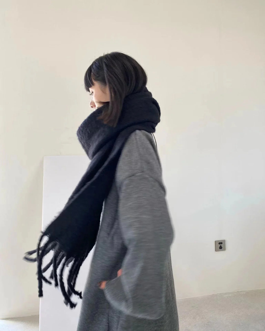 Woman in gray sweater dress wearing a large black fringed scarf in minimalist room