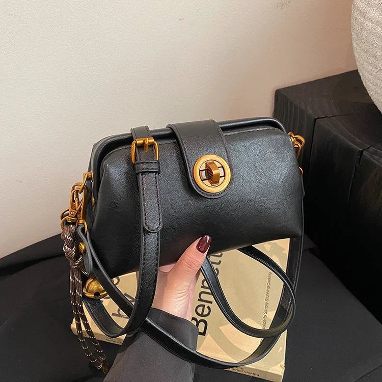 Black leather crossbody bag with gold buckle held by hand over a paper bag on black surface