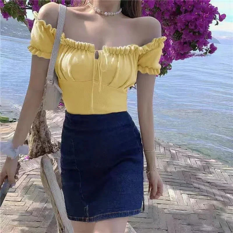 Woman wearing yellow off-shoulder top and dark blue skirt by waterfront with purple flowers