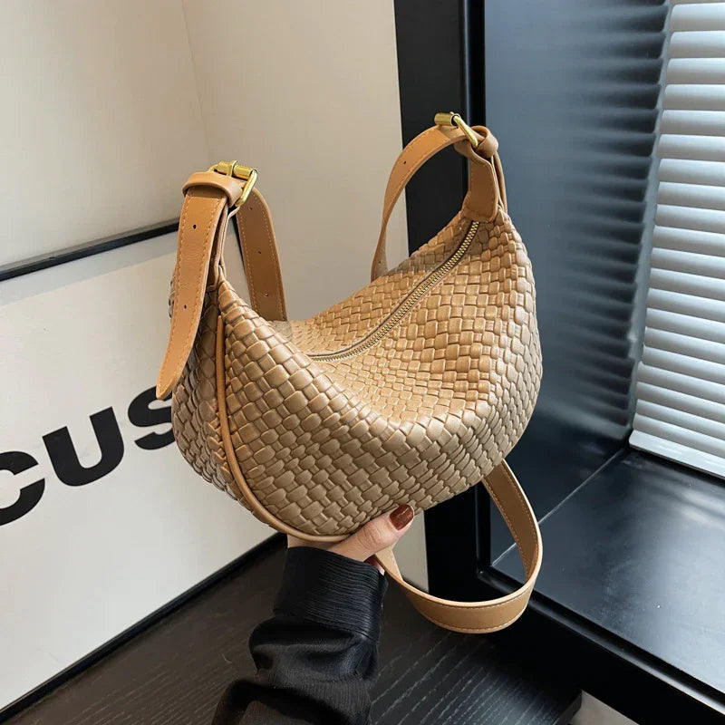 Hand holding a tan woven leather handbag with adjustable straps near a window and black desk