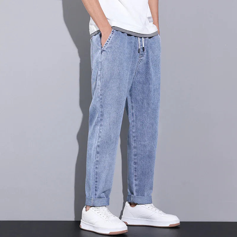Man wearing light blue casual denim jogger pants with elastic cuffs and white sneakers against gray background