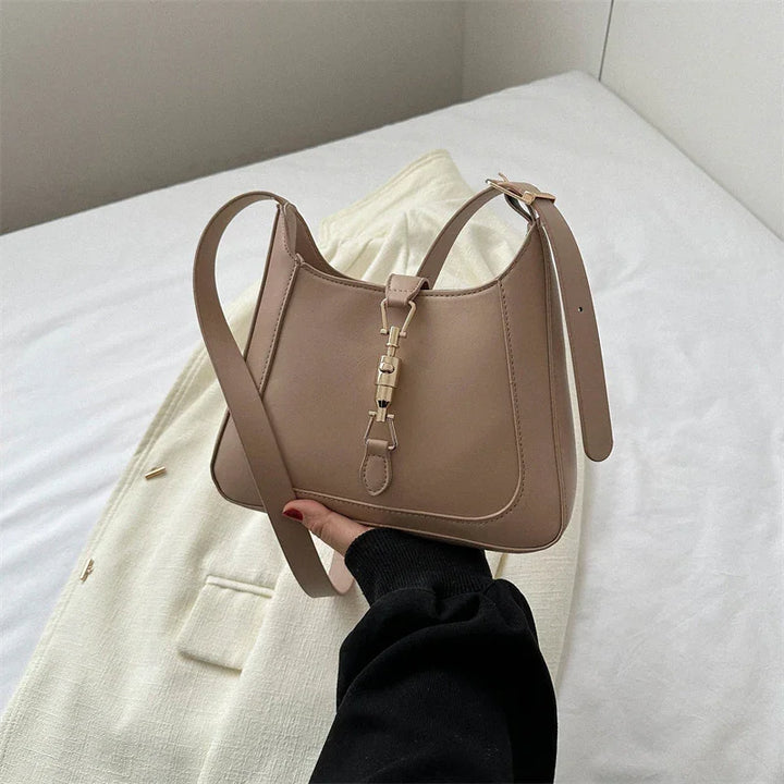 Beige leather handbag with gold clasp and adjustable strap held over white fabric on bed