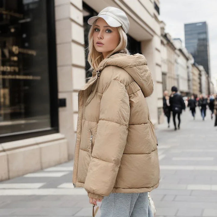Young woman wearing beige puffer jacket and white cap walking on urban street with modern buildings