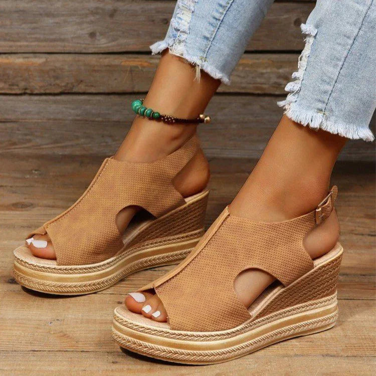 Tan open-toe wedge sandals with cutout design worn with light ripped jeans and green beaded anklet on wooden floor