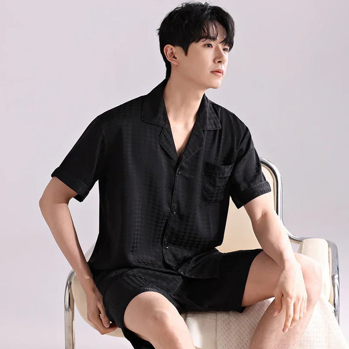 Young man sitting on chair wearing black short-sleeve button-up shirt and shorts loungewear