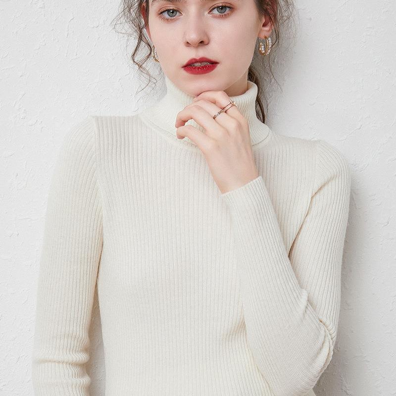 Close-up of woman wearing a cream ribbed turtleneck sweater with gold hoop earrings and rings against white textured wall