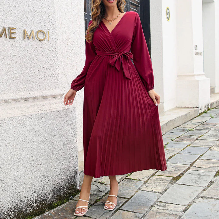 Woman in a long-sleeve burgundy pleated midi dress with a waist tie standing on a stone pathway