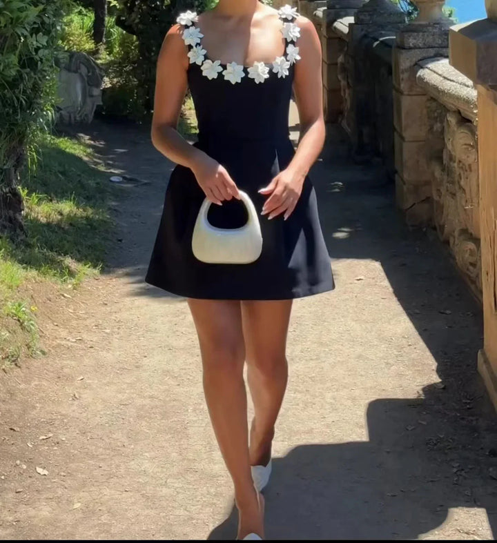 Woman in black mini dress with white flower straps holding white handbag walking outdoors