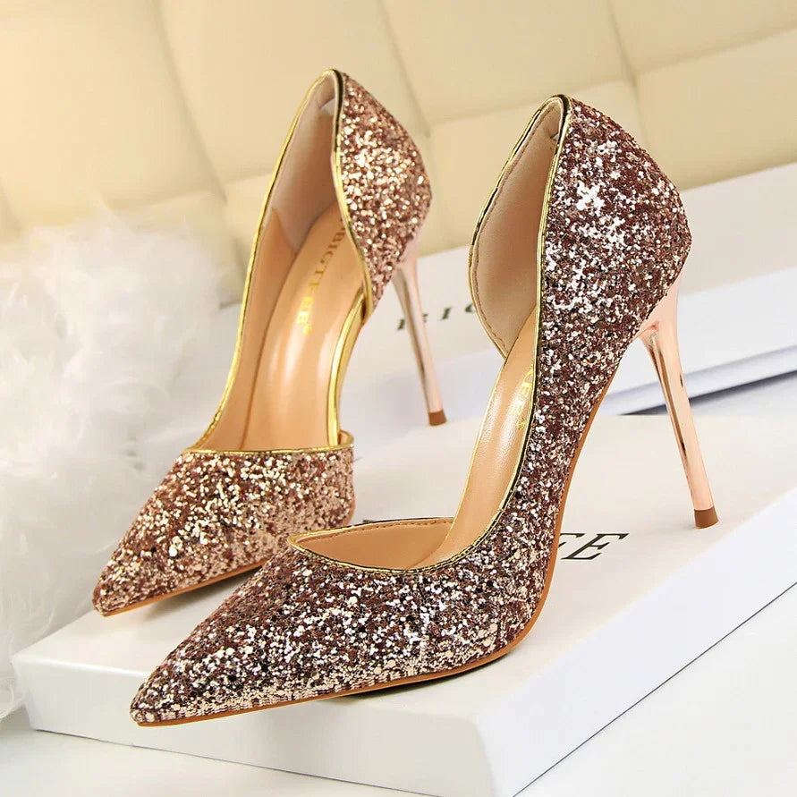 Pair of glittery rose gold high heel shoes with pointed toes on a white surface