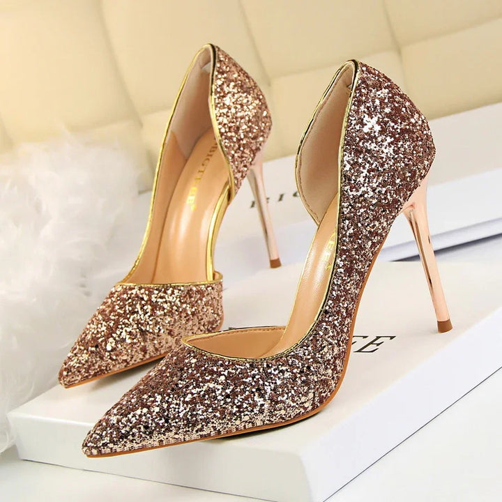 Pair of glittery rose gold high heel shoes with pointed toes on a white surface