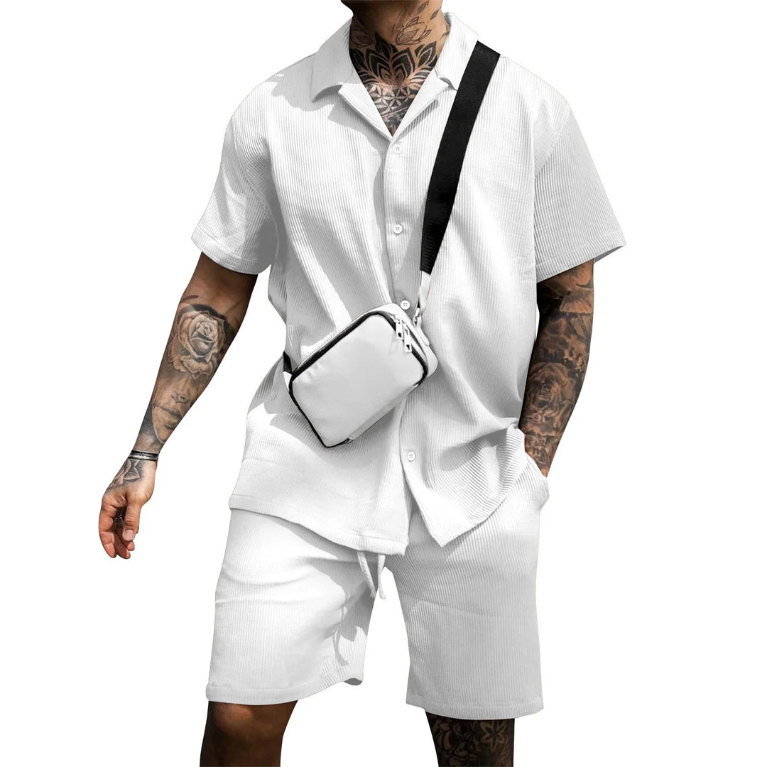 Man wearing white short-sleeve button-up shirt and matching shorts with visible tattoos and a white crossbody bag