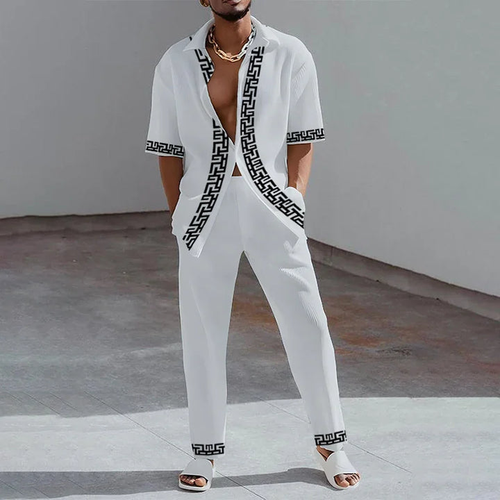 Man wearing stylish white two-piece outfit with black Greek key pattern and white sandals in minimalist setting