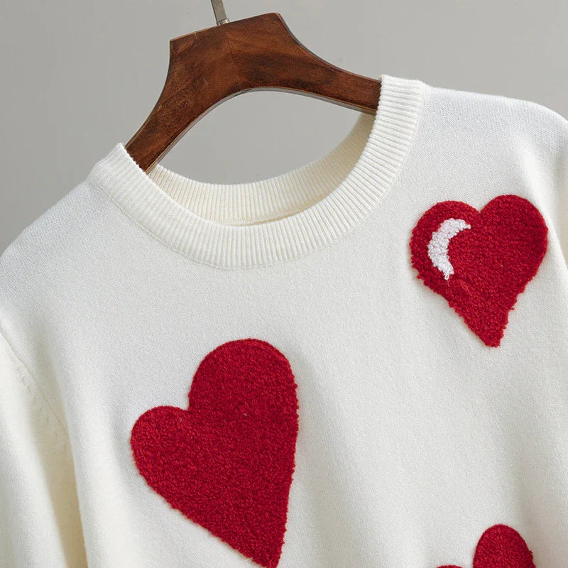 White knit sweater with large red textured heart patches hanging on wooden hanger