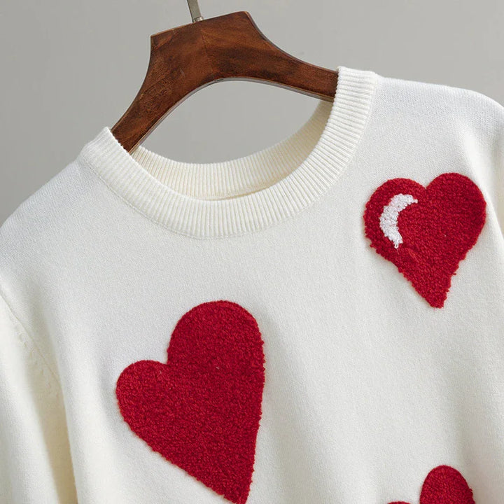 White knit sweater with large red textured heart patches hanging on wooden hanger