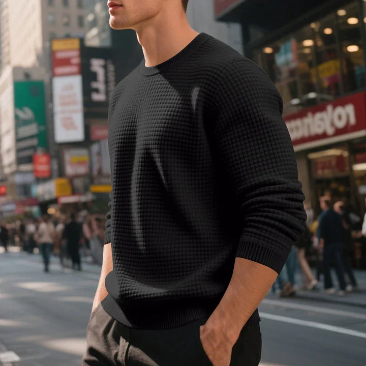 Man wearing black textured sweater with hands in pockets on busy city street