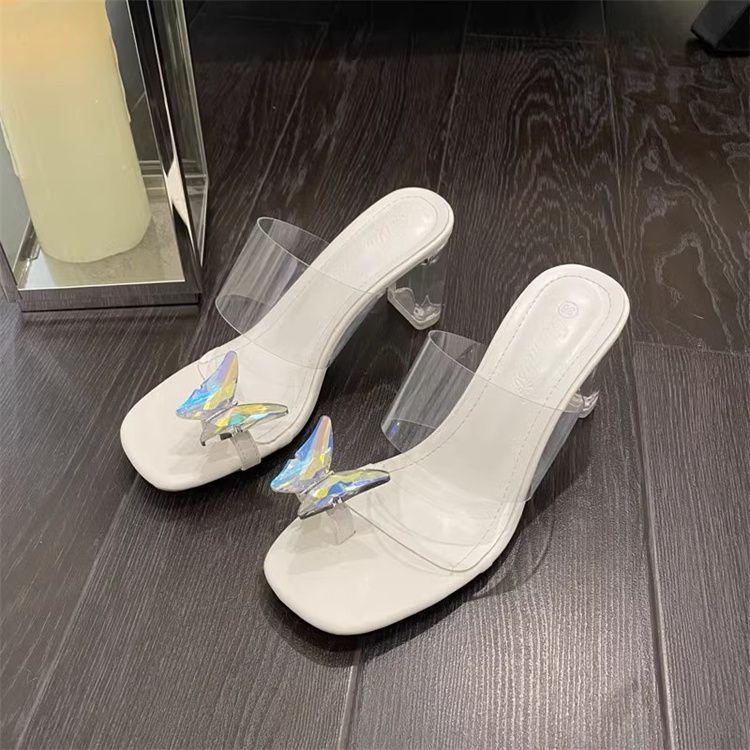 White open-toe mule heels with clear straps and iridescent butterfly embellishments on dark wood floor
