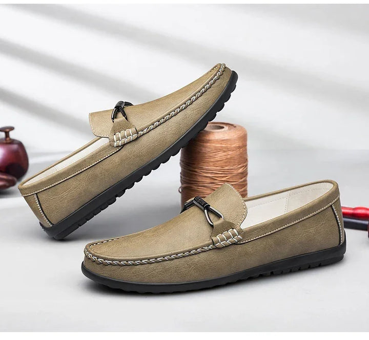 Pair of beige leather loafers with black rubber soles and decorative stitching on a gray surface