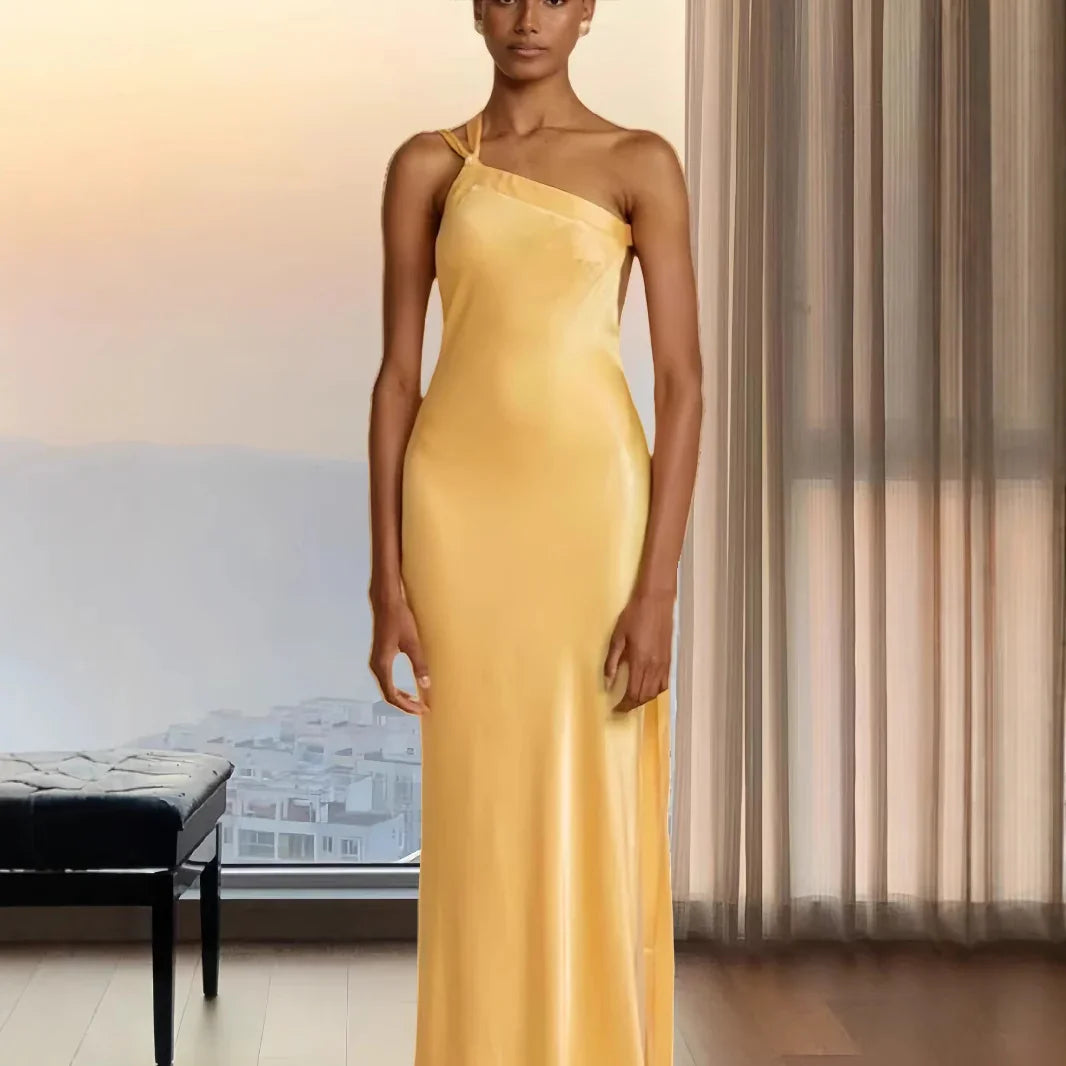 Model wearing elegant yellow satin one-shoulder evening gown standing indoors near window with sheer curtains