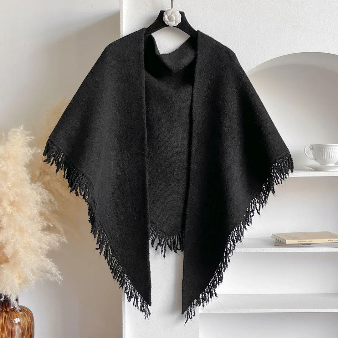 Black wool poncho with fringed edges hanging on a decorative hanger in a modern minimalist room