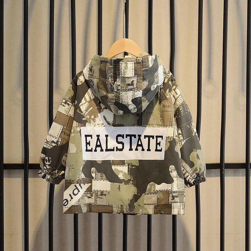 Children's camo hooded jacket with EALSTATE print, hanging on wooden hanger against beige curtain