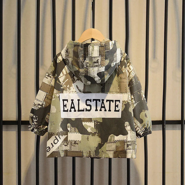 Children's camo hooded jacket with EALSTATE print, hanging on wooden hanger against beige curtain